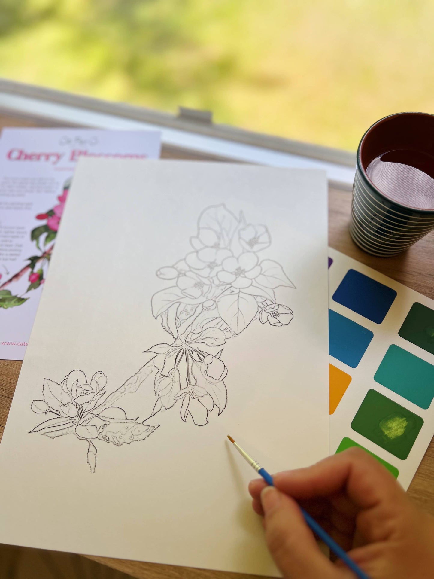 Person painting a floral design on paper with watercolors, surrounded by art supplies.