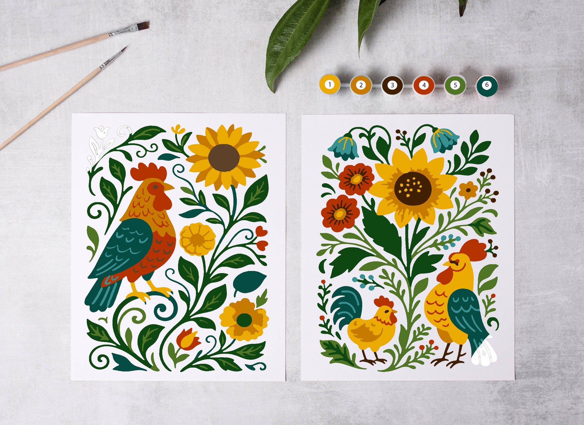 Two colorful illustrations of roosters and flowers on a gray surface with paintbrushes and leaves.