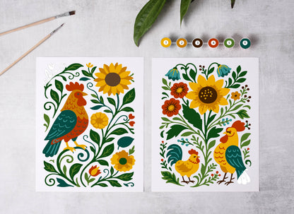 Two colorful illustrations of roosters and flowers on a gray surface with paintbrushes and leaves.