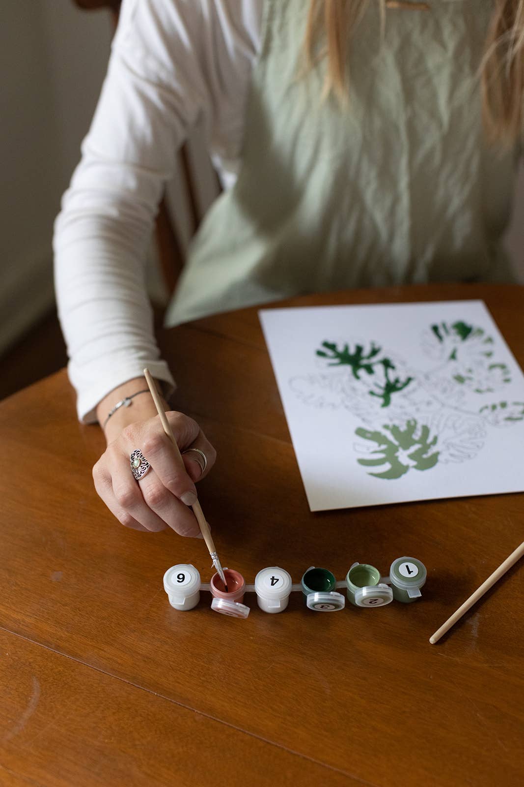 Person painting letters on small round canvases with a paintbrush, next to a sheet of paper with printed letters.