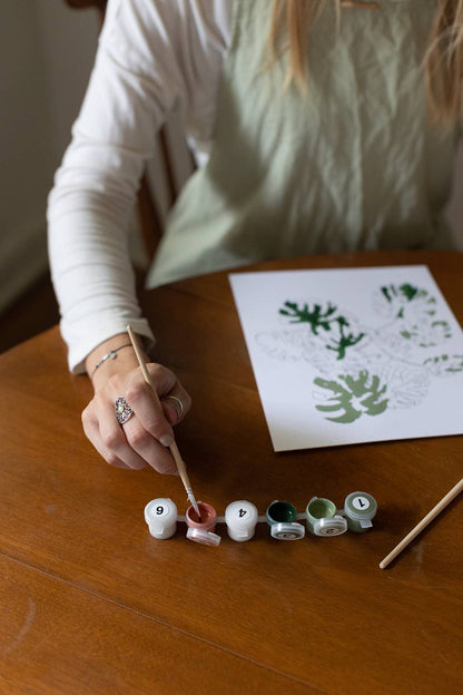 Person painting letters on small round canvases with a paintbrush, next to a sheet of paper with printed letters.