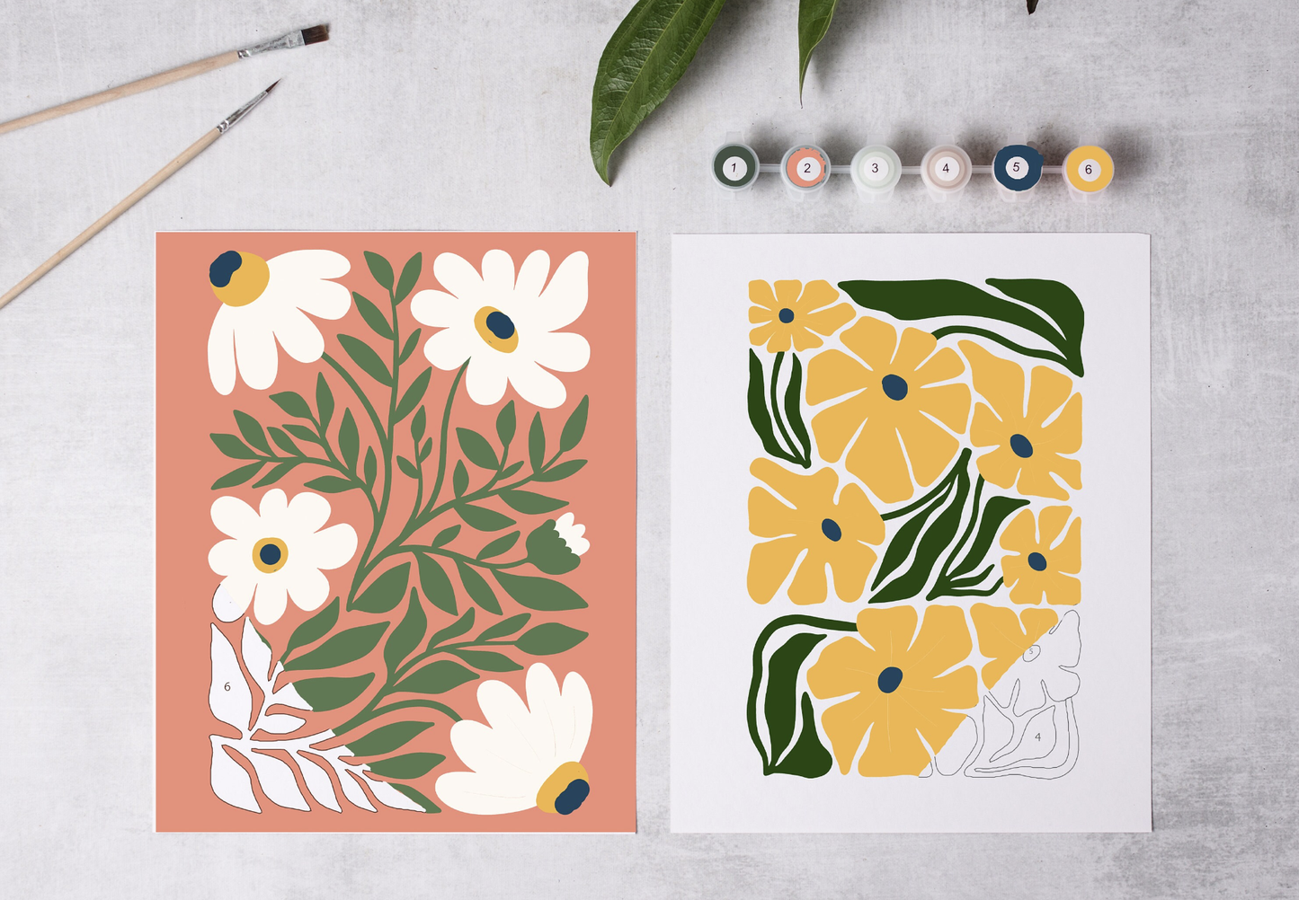 Two floral designs on paper with paintbrushes and paint tubes in the background.