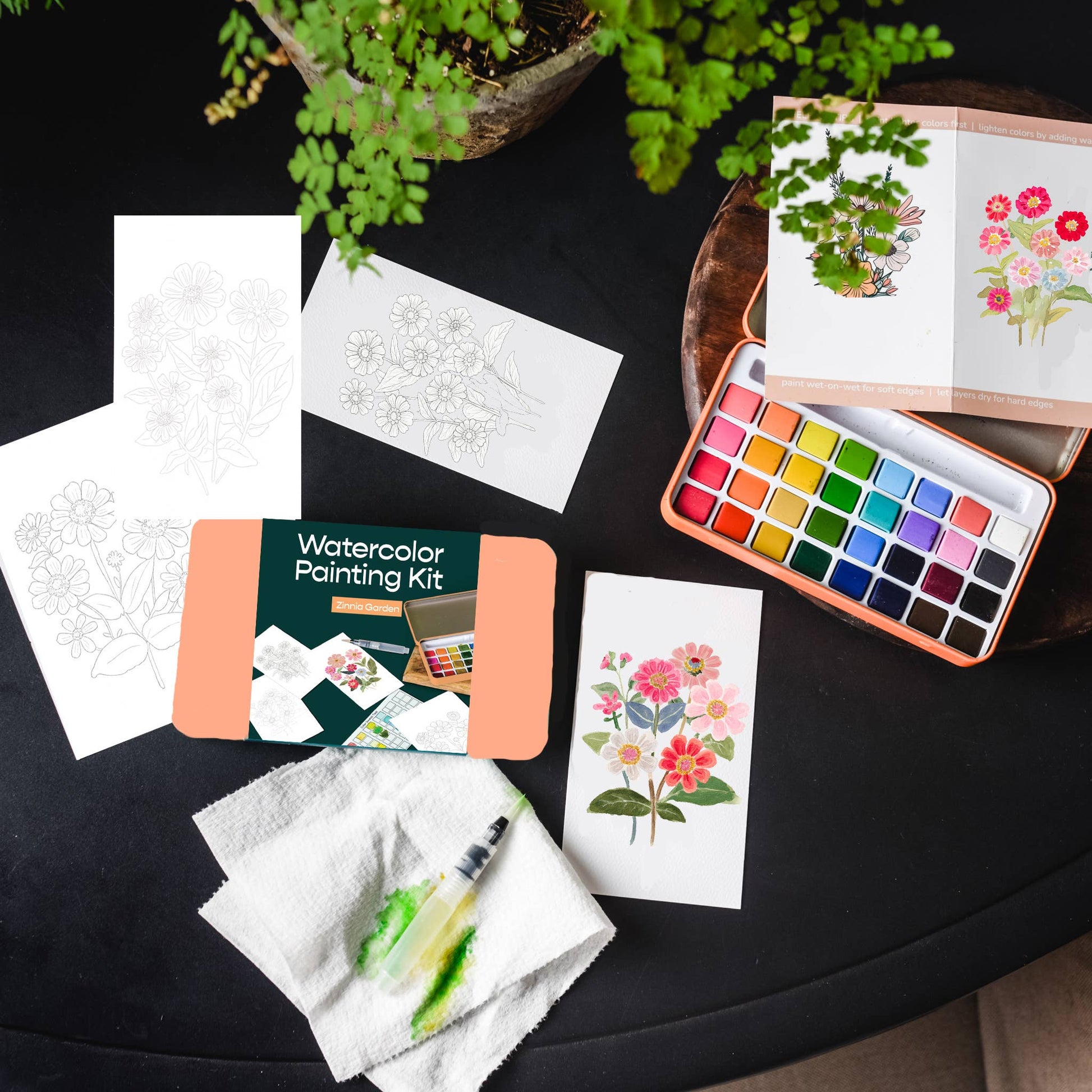 Watercolor painting kit with watercolor paper, paint set, and a plant on a dark surface.