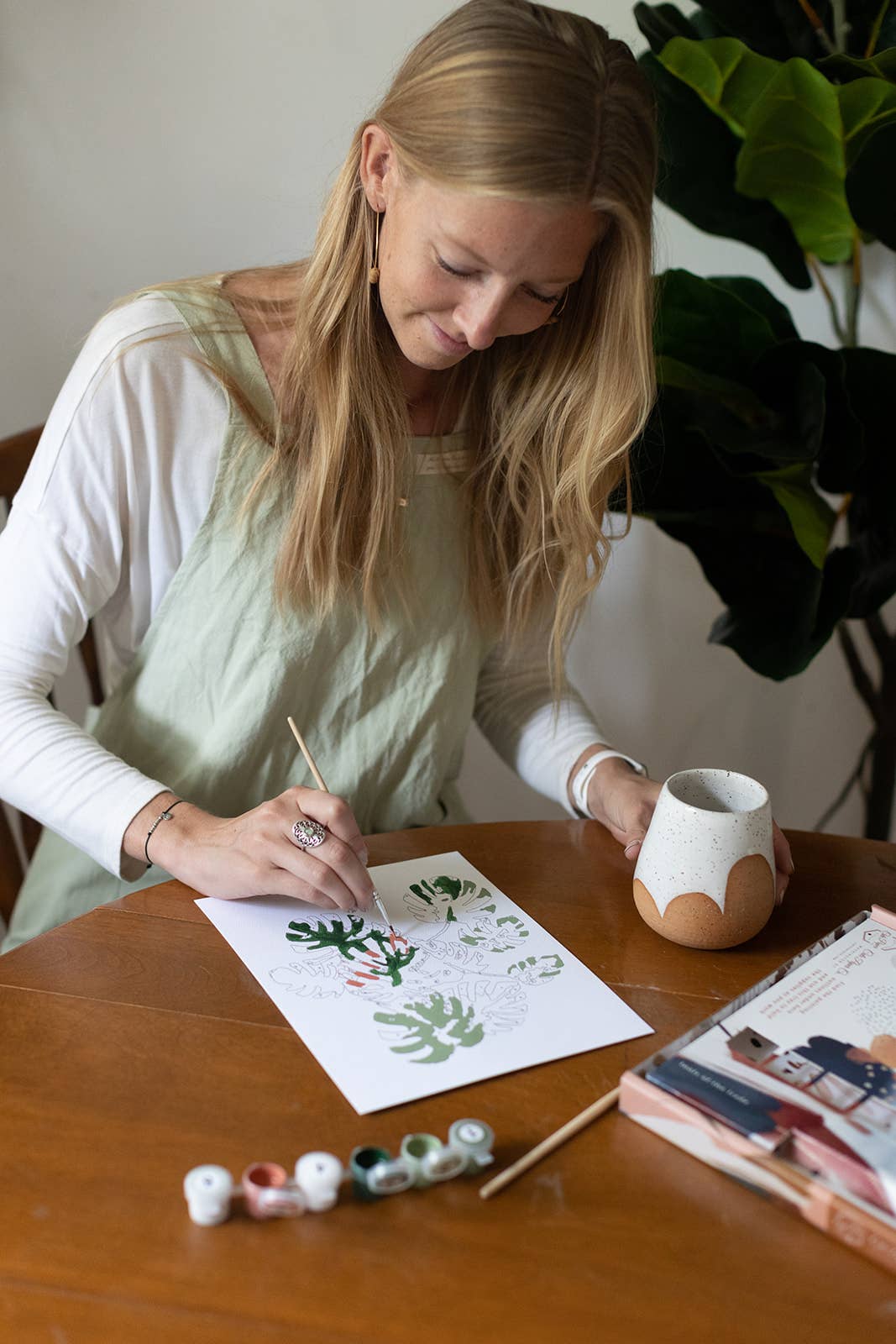 Woman painting a design on paper with a cup and art supplies on a table.