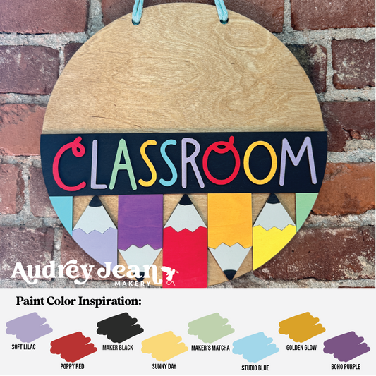 A round wooden classroom sign hanging against a brick wall, with colorful painted pencil shapes and the word 'CLASSROOM' across the center. A watermark reading Audrey Jean's Makers appears at the lower left, and a color inspiration chart is shown along the bottom.