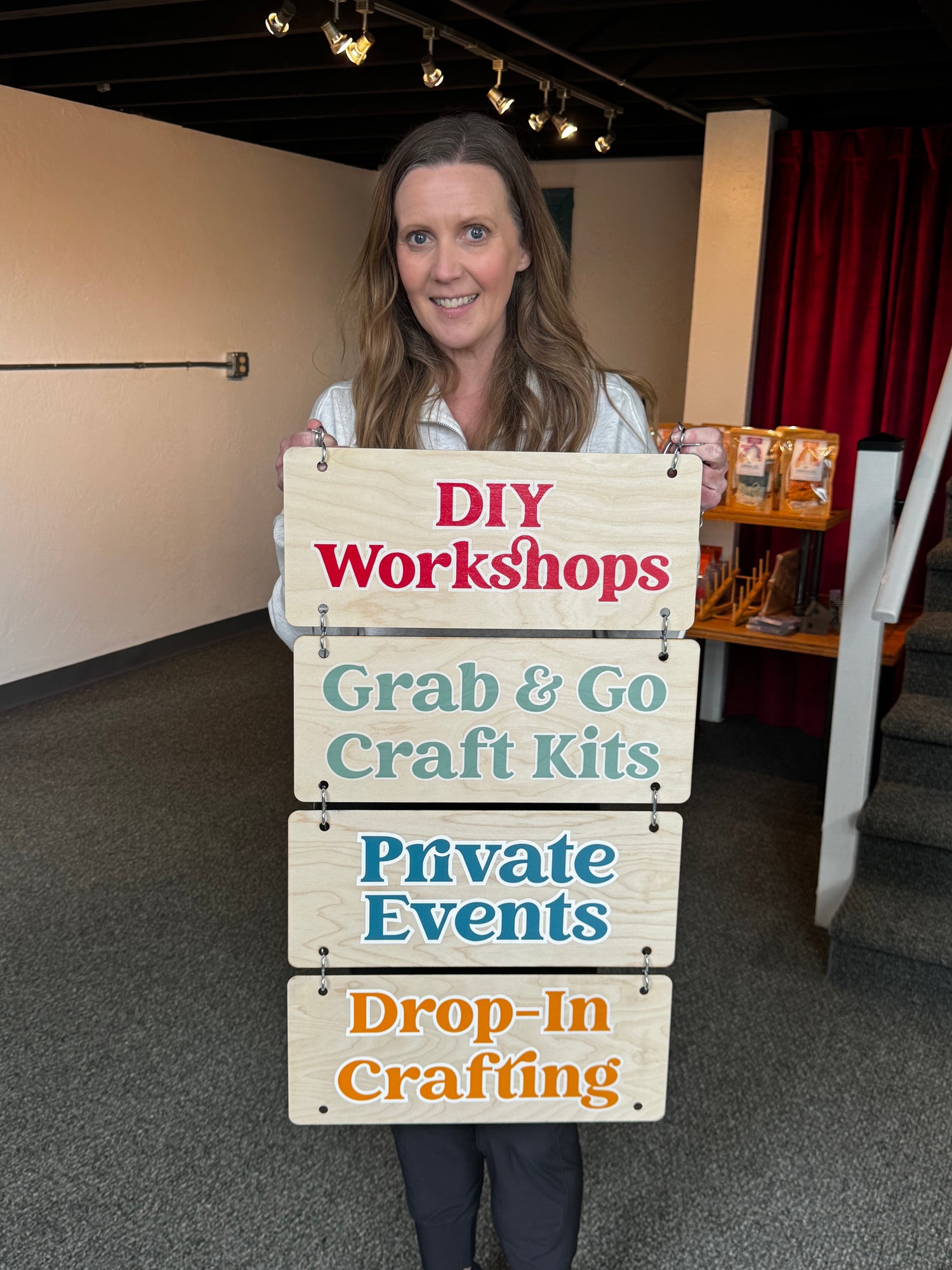 Woman holding a sign with text about DIY workshops and craft offerings in an indoor setting.