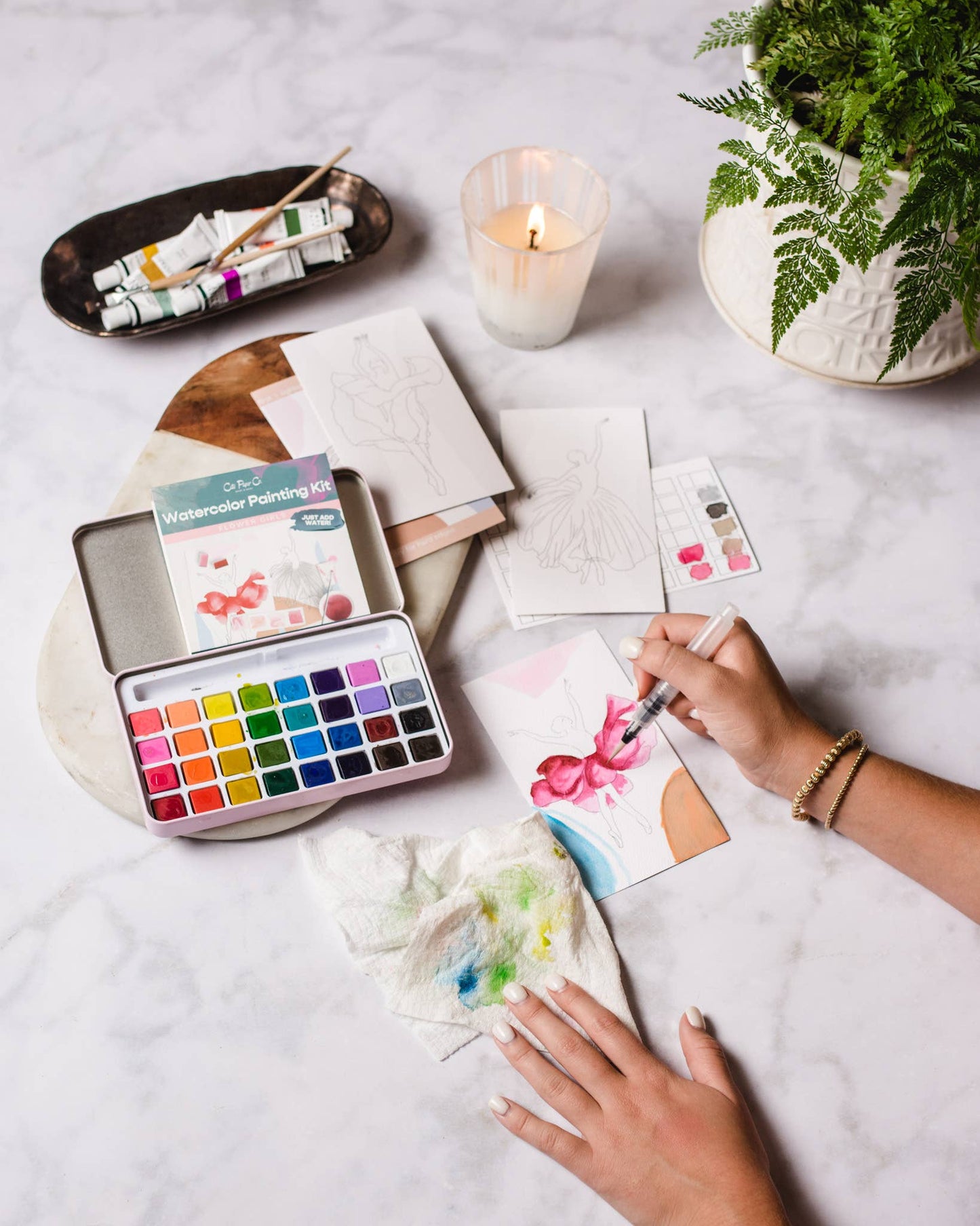 Person painting with watercolors on a table with art supplies and a candle.
