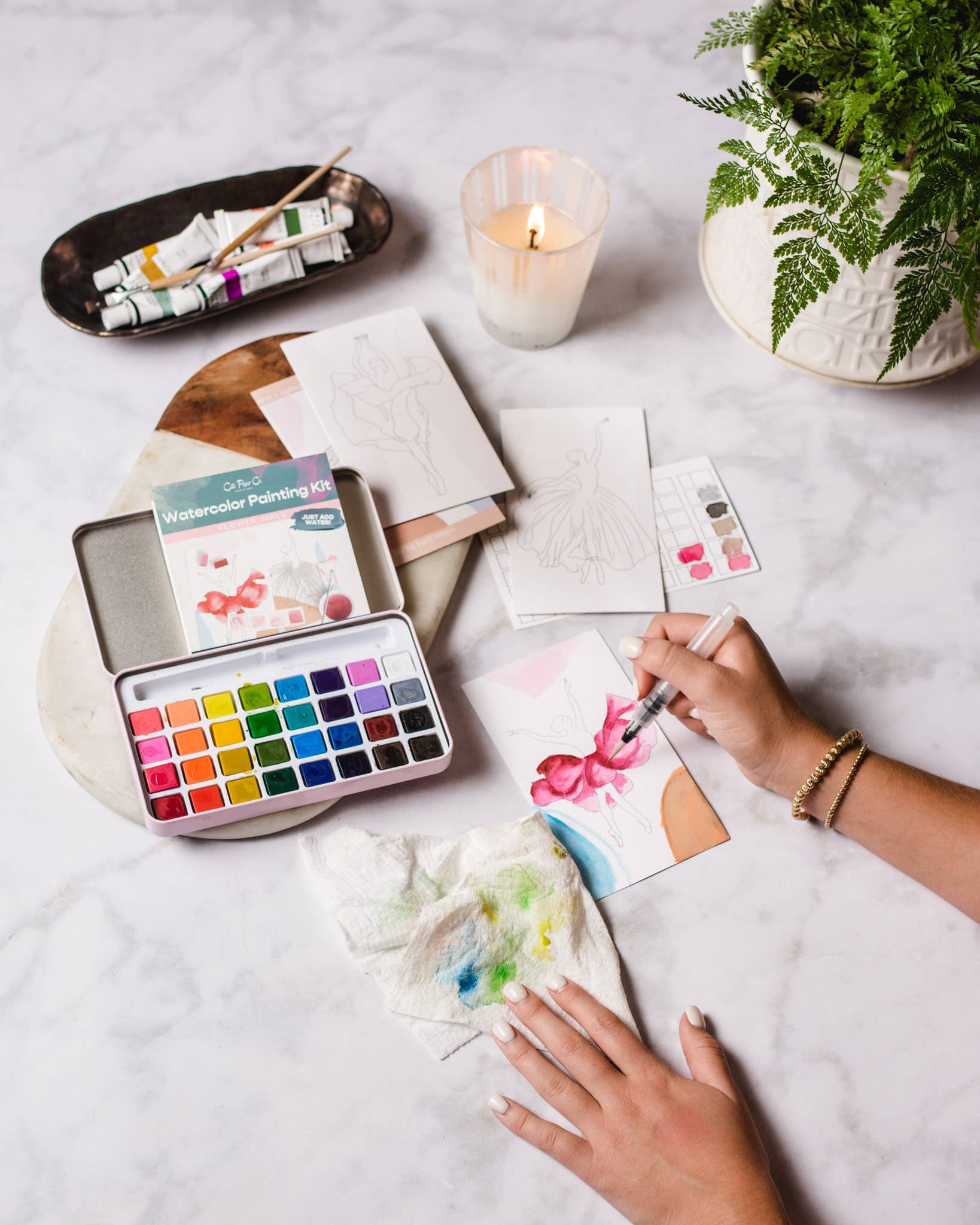 Person painting with watercolors on a table with art supplies and a candle.