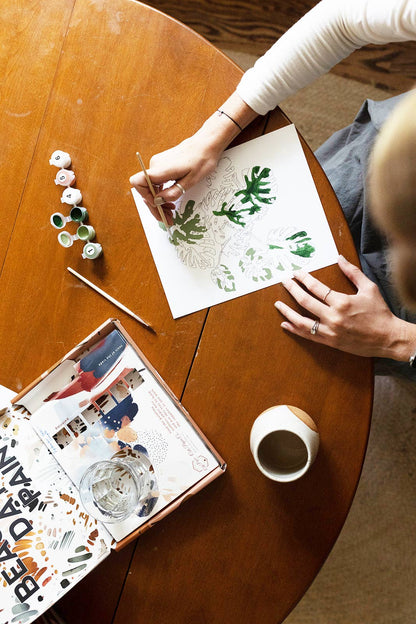 Person painting on paper with a cup of coffee and art supplies on a wooden table