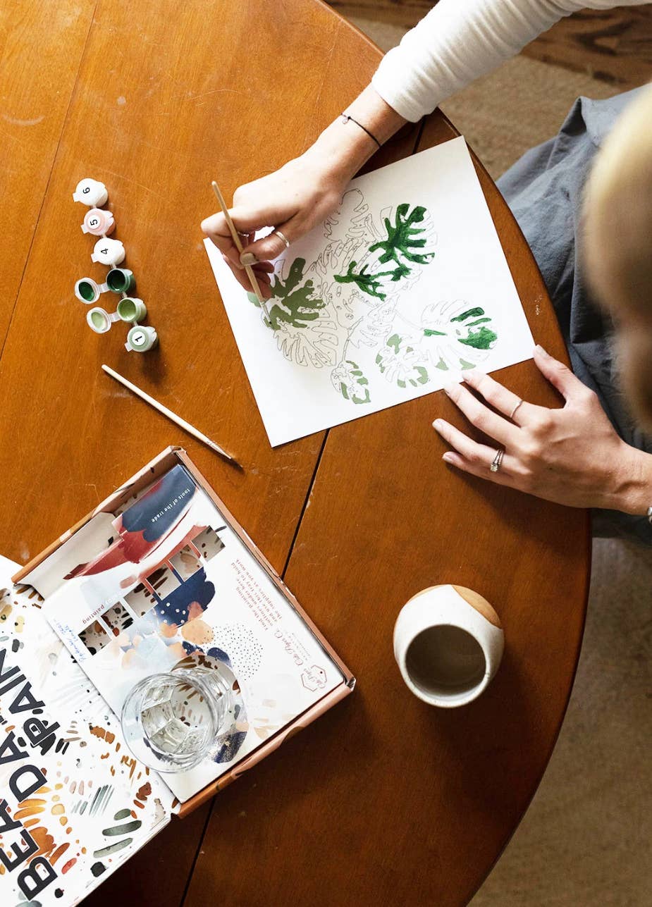 Person painting on paper with a cup of coffee and art supplies on a wooden table