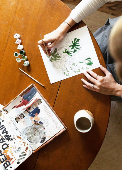 Person painting on paper with a cup of coffee and art supplies on a wooden table